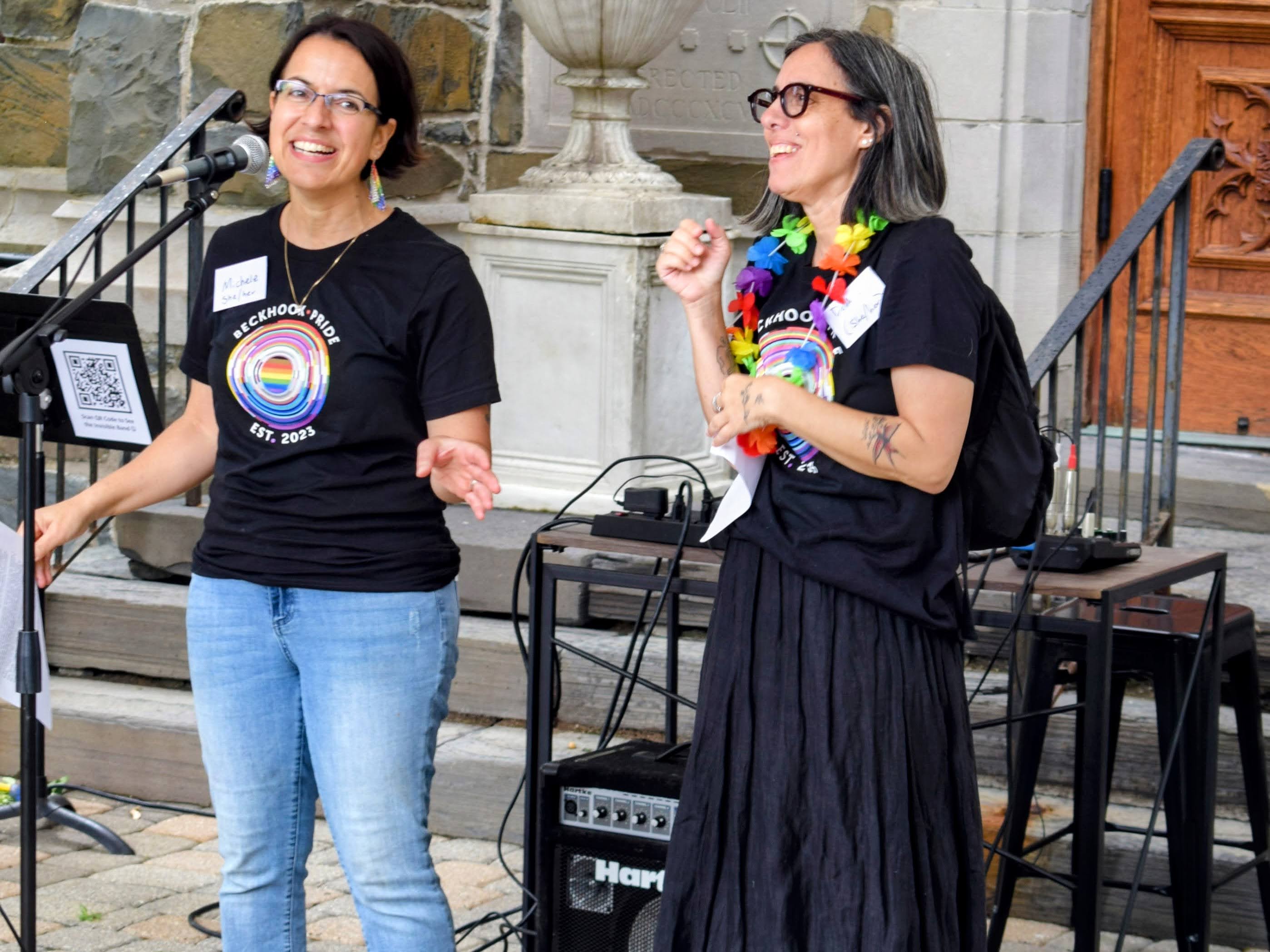 Two women behind a microphone, one with a plastic lei